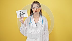 Young beautiful hispanic woman doctor standing with serious expression holding we need you paper over isolated yellow background