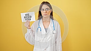 Young beautiful hispanic woman doctor standing with serious expression holding we need you paper over isolated yellow background