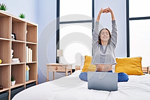 Young beautiful hispanic woman doing online stretching exercise sitting on bed at bedroom