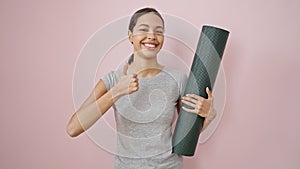 Young beautiful hispanic woman doing thumbs up wearing sportswear holding yoga mat over isolated pink background