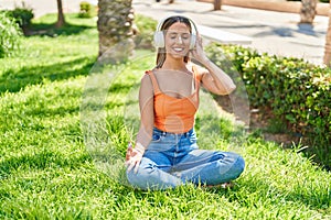Young beautiful hispanic woman doing yoga exercise using headphones at park