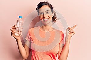 Young beautiful hispanic woman drinking bottle of water smiling happy pointing with hand and finger to the side