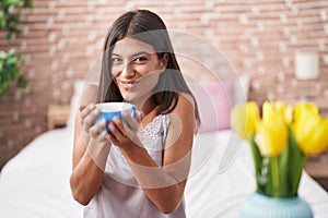 Young beautiful hispanic woman drinking cup of coffee sitting on bed at bedroom