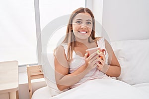 Young beautiful hispanic woman drinking cup of coffee sitting on bed at bedroom