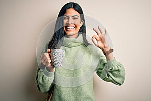 Young beautiful hispanic woman drinking a cup of hot coffee over isolated background doing ok sign with fingers, excellent symbol
