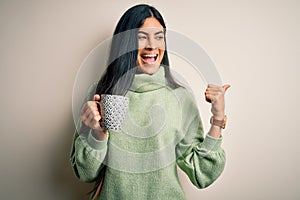 Young beautiful hispanic woman drinking a cup of hot coffee over isolated background pointing and showing with thumb up to the