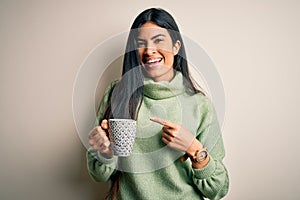 Young beautiful hispanic woman drinking a cup of hot coffee over isolated background very happy pointing with hand and finger