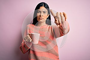 Young beautiful hispanic woman drinking a cup of hot coffee over pink isolated background pointing with finger to the camera and