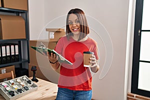 Young beautiful hispanic woman ecommerce business worker holding checklist drinking coffee at office