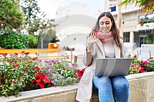 Young beautiful hispanic woman having video call sitting on bench at park