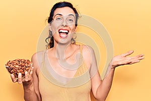 Young beautiful hispanic woman holding bowl with almonds celebrating achievement with happy smile and winner expression with