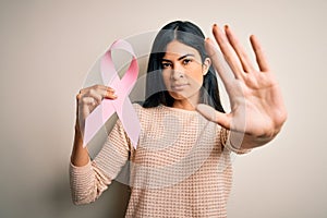 Young beautiful hispanic woman holding breast cancer awareness pink ribbon with open hand doing stop sign with serious and