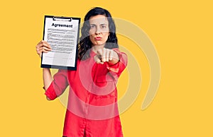 Young beautiful hispanic woman holding clipboard with agreement document pointing with finger to the camera and to you, confident