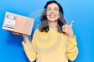 Young beautiful hispanic woman holding delivery package smiling happy and positive, thumb up doing excellent and approval sign