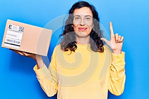 Young beautiful hispanic woman holding delivery package smiling with an idea or question pointing finger with happy face, number