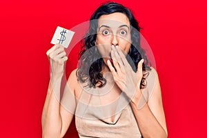 Young beautiful hispanic woman holding dollar symbol reminder paper covering mouth with hand, shocked and afraid for mistake