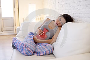 Young beautiful hispanic woman holding hot water bottle against belly suffering menstrual period pain