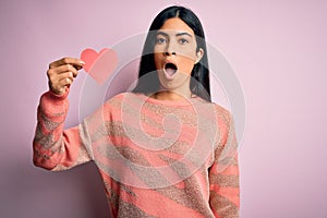 Young beautiful hispanic woman holding romantic heart paper shape over pink background scared in shock with a surprise face,