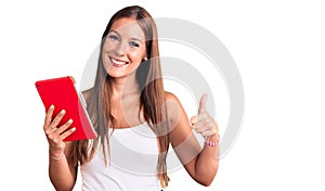 Young beautiful hispanic woman holding touchpad smiling happy and positive, thumb up doing excellent and approval sign