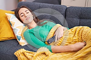 Young beautiful hispanic woman holding tv remote control lying on sofa sleeping at home