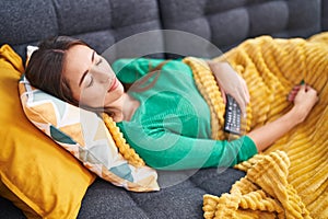 Young beautiful hispanic woman holding tv remote control lying on sofa sleeping at home