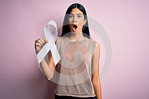 Young beautiful hispanic woman holding white ribbon as stop violence and lung cancer symbol scared in shock with a surprise face,