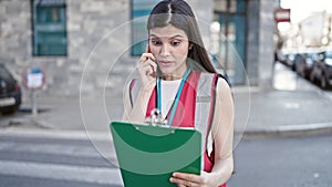 Young beautiful hispanic woman interviewer talking on smartphone reading document at street