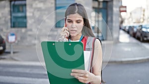 Young beautiful hispanic woman interviewer talking on smartphone reading document at street