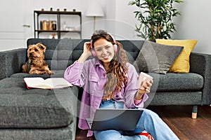 Young beautiful hispanic woman listening to music sitting on floor with dog at home