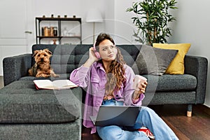 Young beautiful hispanic woman listening to music sitting on floor with dog at home