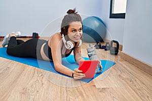Young beautiful hispanic woman lying on yoga mat using touchpad at sport center
