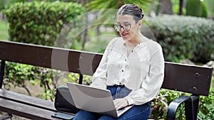 Young beautiful hispanic woman on an online meeting with laptop sitting on a bench at the park