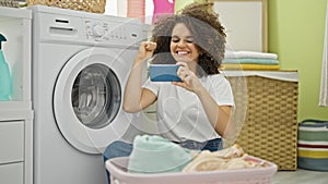 Young beautiful hispanic woman playing video game waiting for washing machine at laundry room