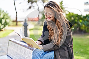 Young beautiful hispanic woman reading book sitting on bench at park