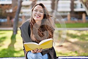 Young beautiful hispanic woman reading book sitting on bench at park