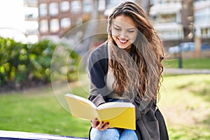 Young beautiful hispanic woman reading book sitting on bench at park