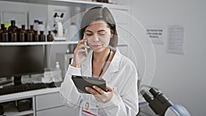 Young beautiful hispanic woman scientist using touchpad talking on smartphone at laboratory