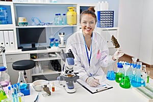 Young beautiful hispanic woman scientist writing report holding marijuana at laboratory