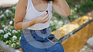 Young beautiful hispanic woman sitting on bench holding earphones and smartphone at park