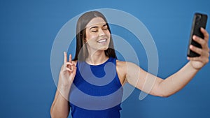 Young beautiful hispanic woman smiling confident making selfie by the smartphone over isolated blue background