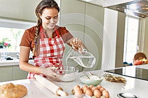 Young beautiful hispanic woman smiling confident pouring water on bowl with flour at the kitchen