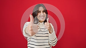 Young beautiful hispanic woman smiling with thumbs up over isolated red background