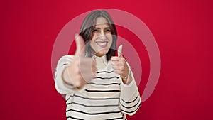 Young beautiful hispanic woman smiling with thumbs up over isolated red background