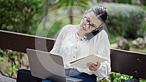 Young beautiful hispanic woman speaking on the phone writing on a notebook sitting on a bench at the park