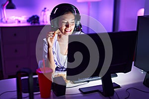 Young beautiful hispanic woman streamer playing video game eating chips potatoes at gaming room