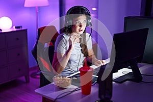 Young beautiful hispanic woman streamer playing video game eating chips potatoes at gaming room