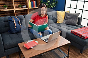 Young beautiful hispanic woman student sitting on sofa studying at home