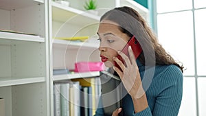 Young beautiful hispanic woman student speaking on the phone looking for a book at university classroom