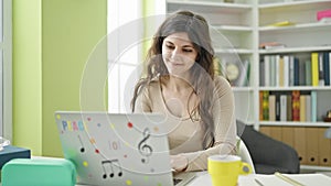 Young beautiful hispanic woman student using laptop drinking coffee at library university