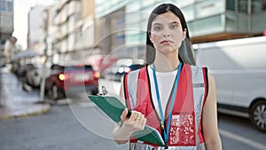 Young beautiful hispanic woman survey interviewer holding clipboard standing with serious expression at street